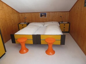 A small, single-bed room with wooden walls, a nightstand with a lamp, and a painting on the wall.