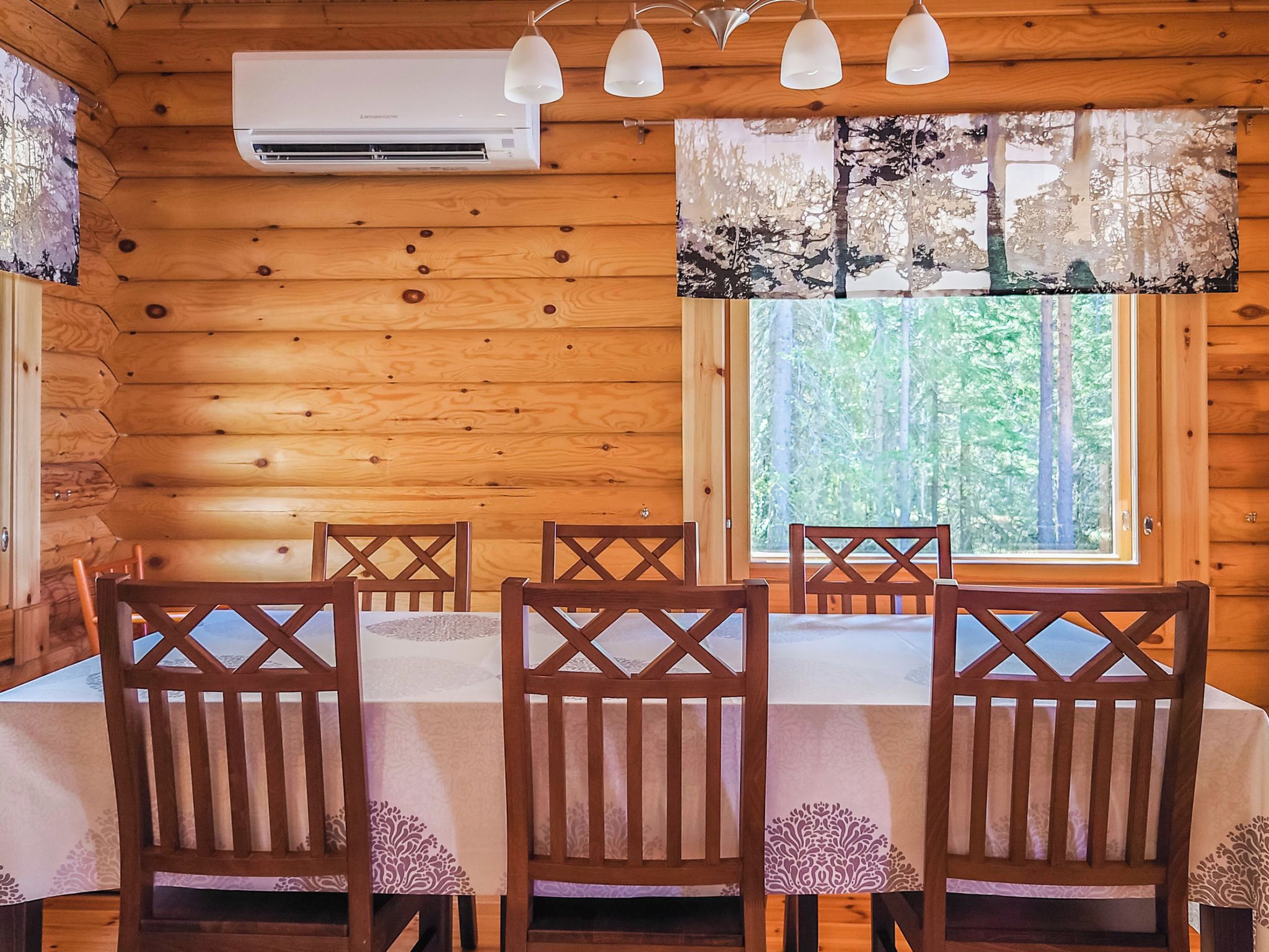 Puinen mökki, jonka sisustuksessa on ruokapöytä neljälle, puiset tuolit, ilmastointilaite ja ikkuna, josta avautuu näkymä puihin.