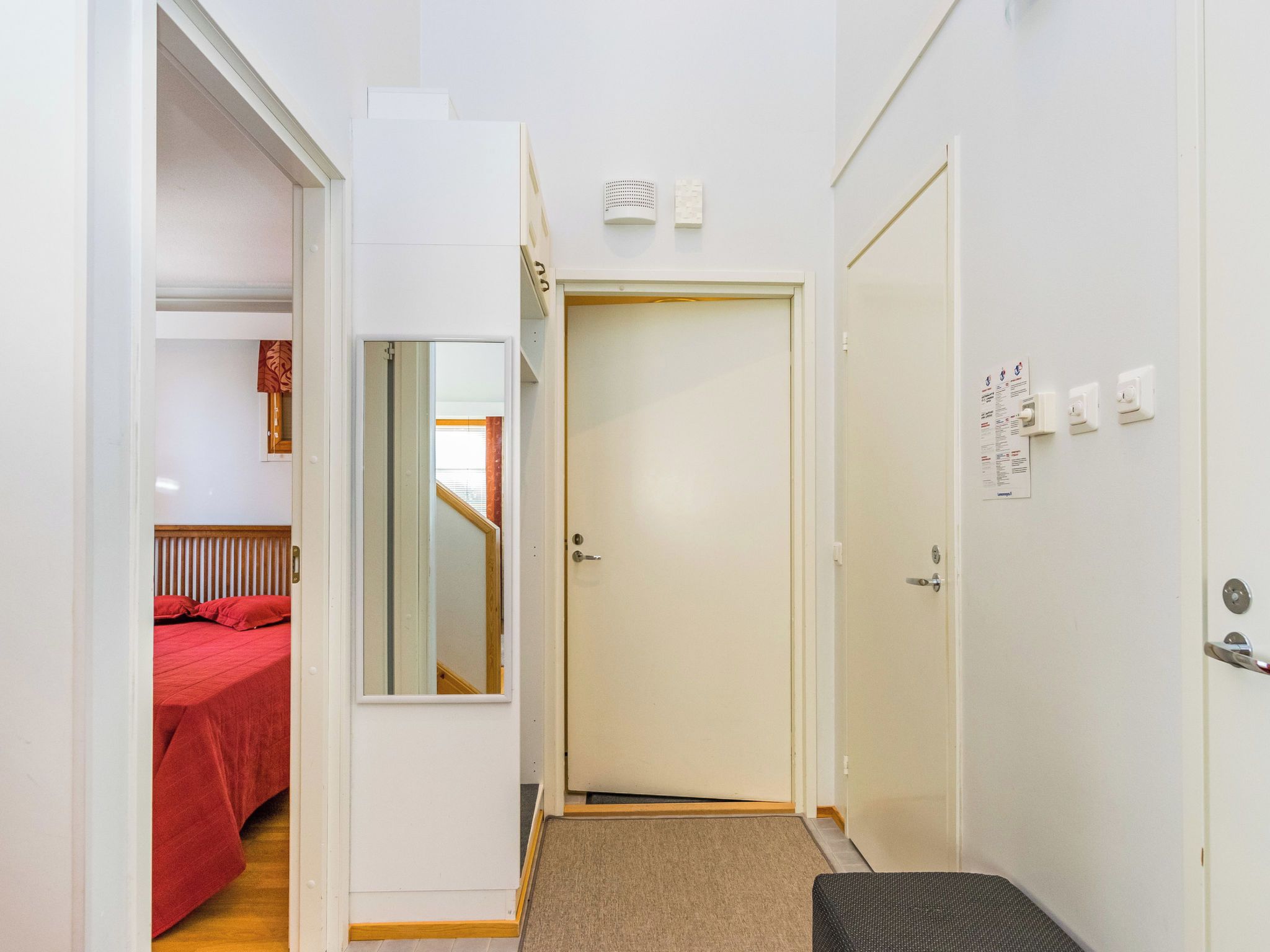 En smal hall med en dörr som leder till ett sovrum med ett rött sängöverkast.