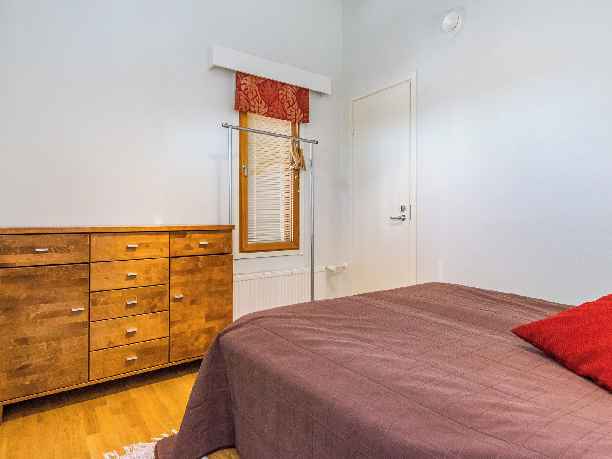 A bedroom with a wooden dresser, a bed with a red pillow, a window with blinds, and a white door.