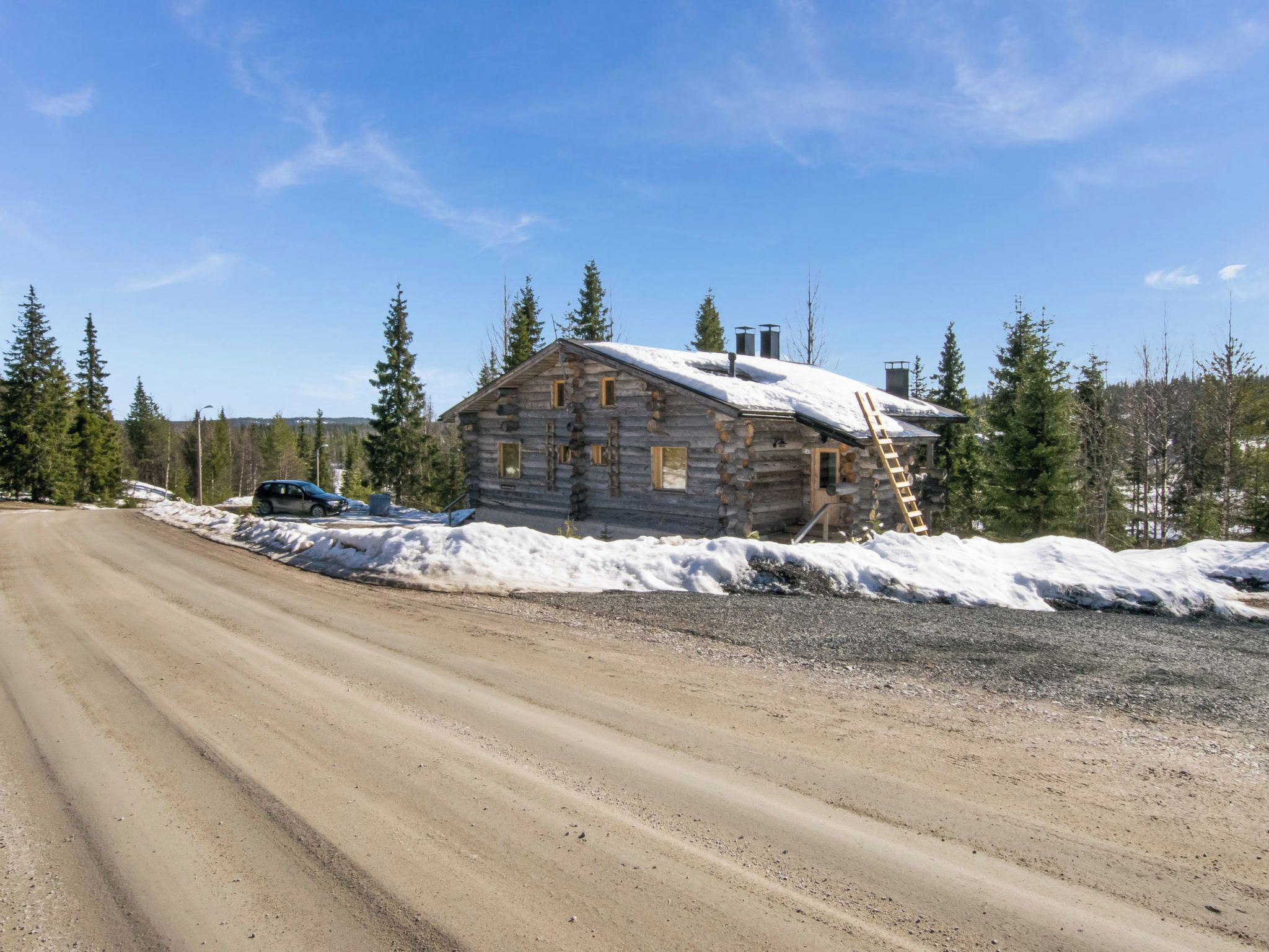 A log cabin with a dirt road in front, surrounded by snow and trees.