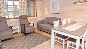 Ein gemütliches Wohnzimmer mit einer Couch, zwei Sesseln, einem Esstisch mit Stühlen, einem gemusterten Teppich und einem großen Fenster mit Jalousien.