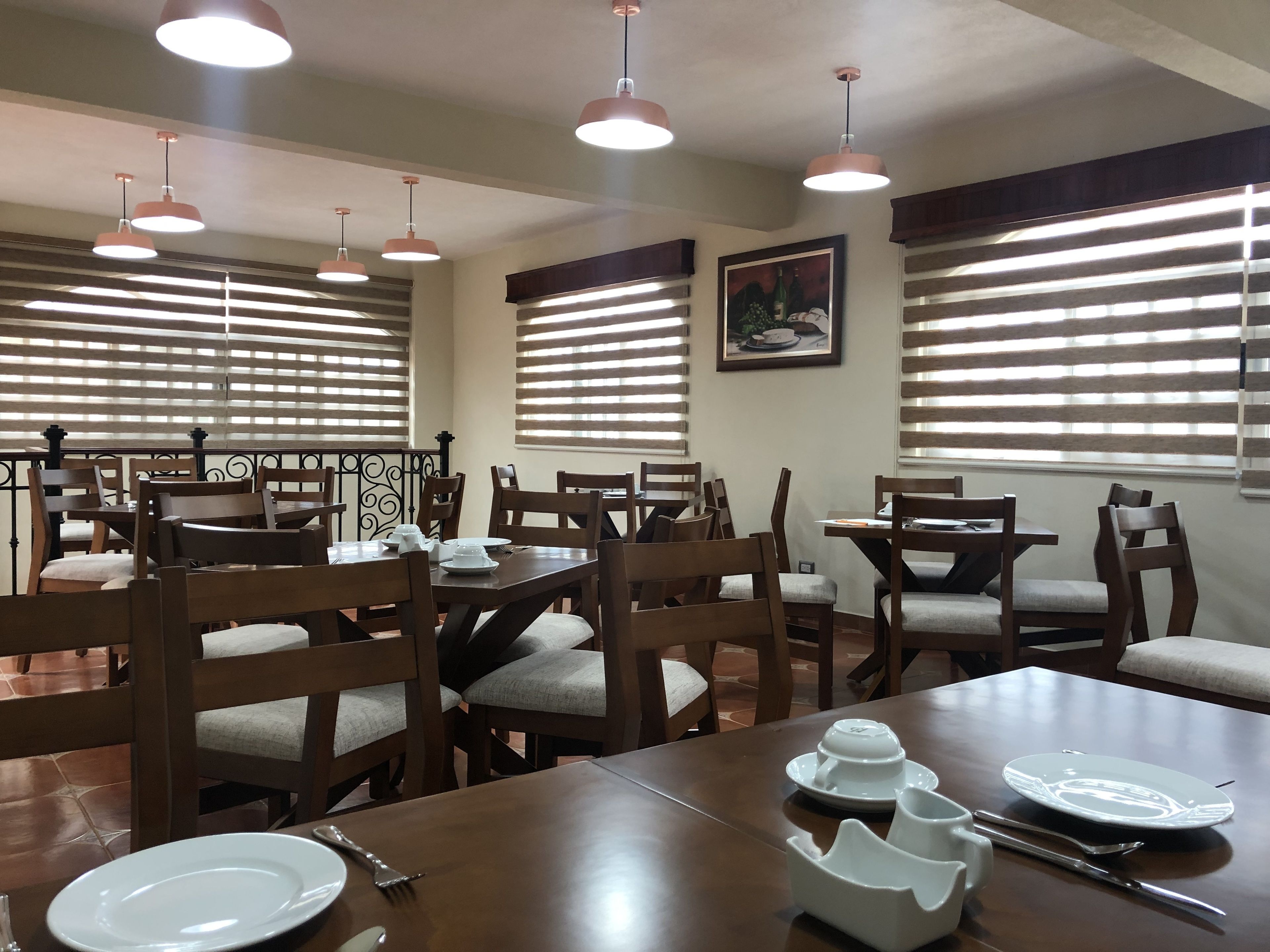 Un comedor con mesas y sillas de madera, iluminación colgante y ventanas amplias con persianas.