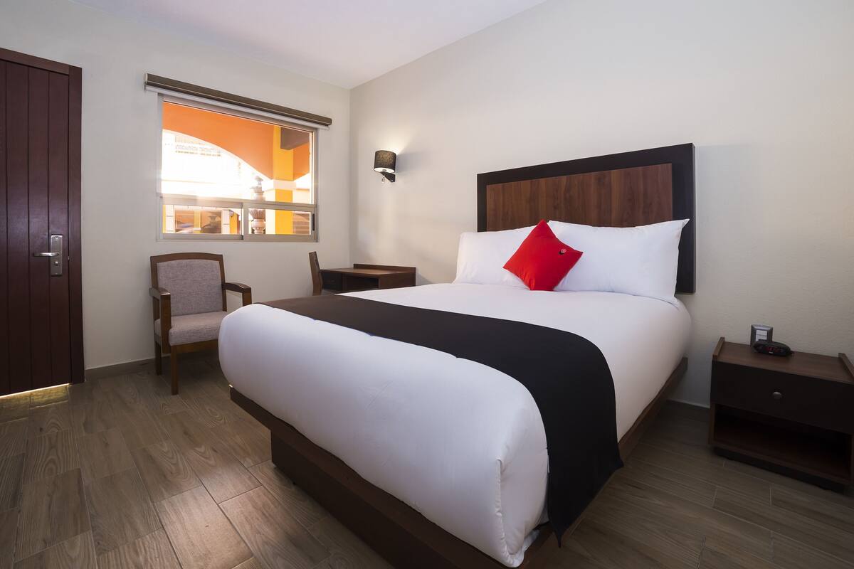 Une chambre d’hôtel avec un grand lit, un bureau, une chaise, une table de chevet et une fenêtre avec des stores.