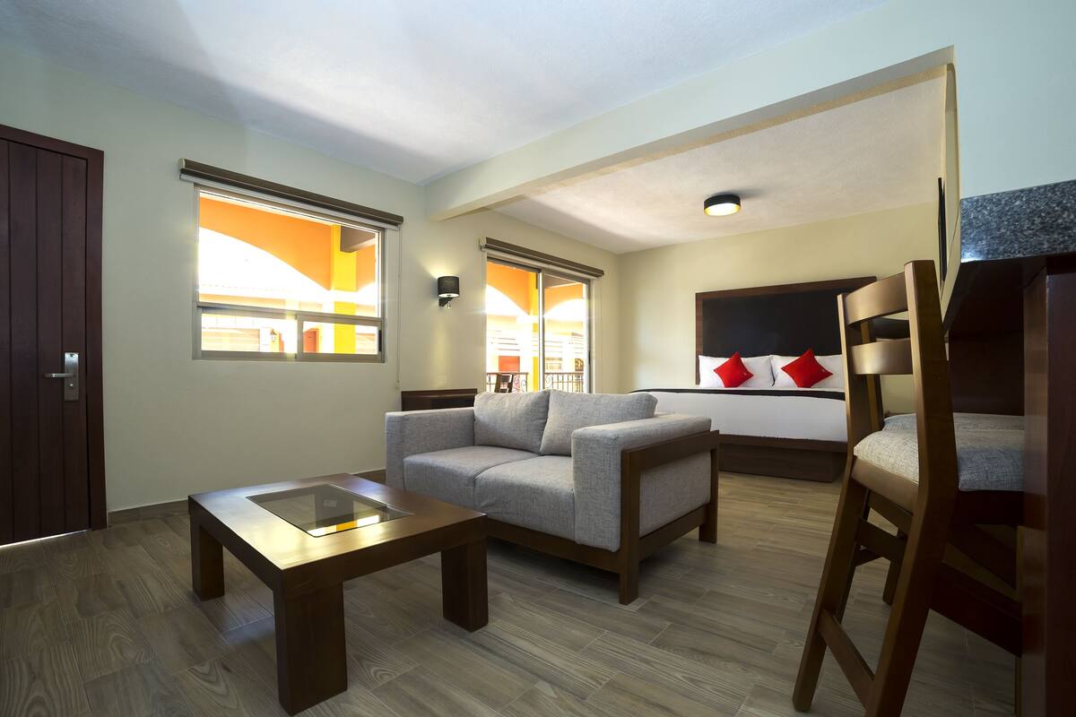 Une chambre d’hôtel avec un canapé, une table basse en bois, un lit avec des oreillers rouges et blancs, et une chaise en bois.