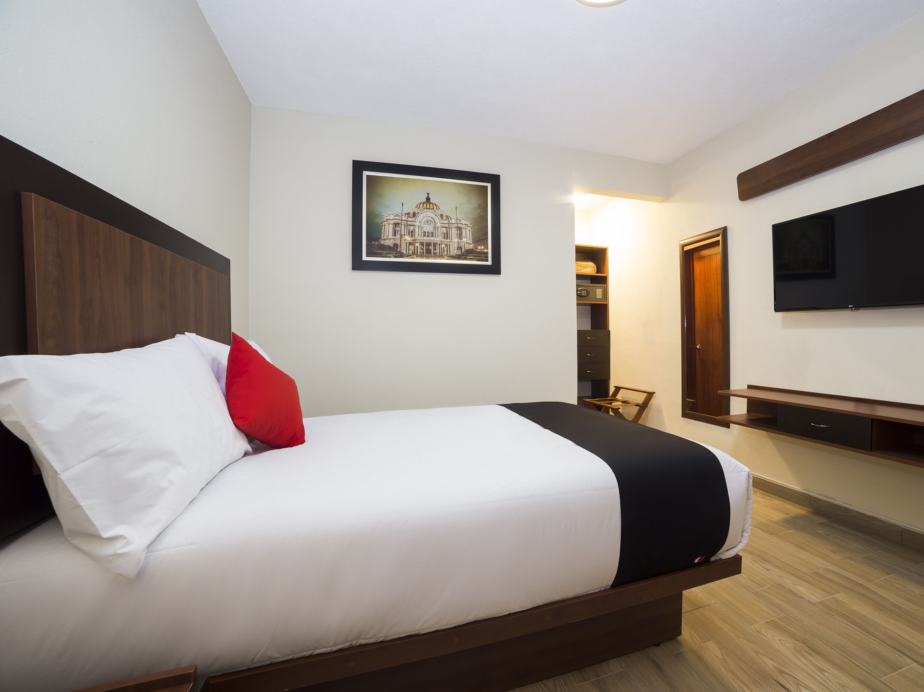 Una cama bien tendida con ropa de cama blanca y negra, un cojín rojo y un cuadro en la pared.