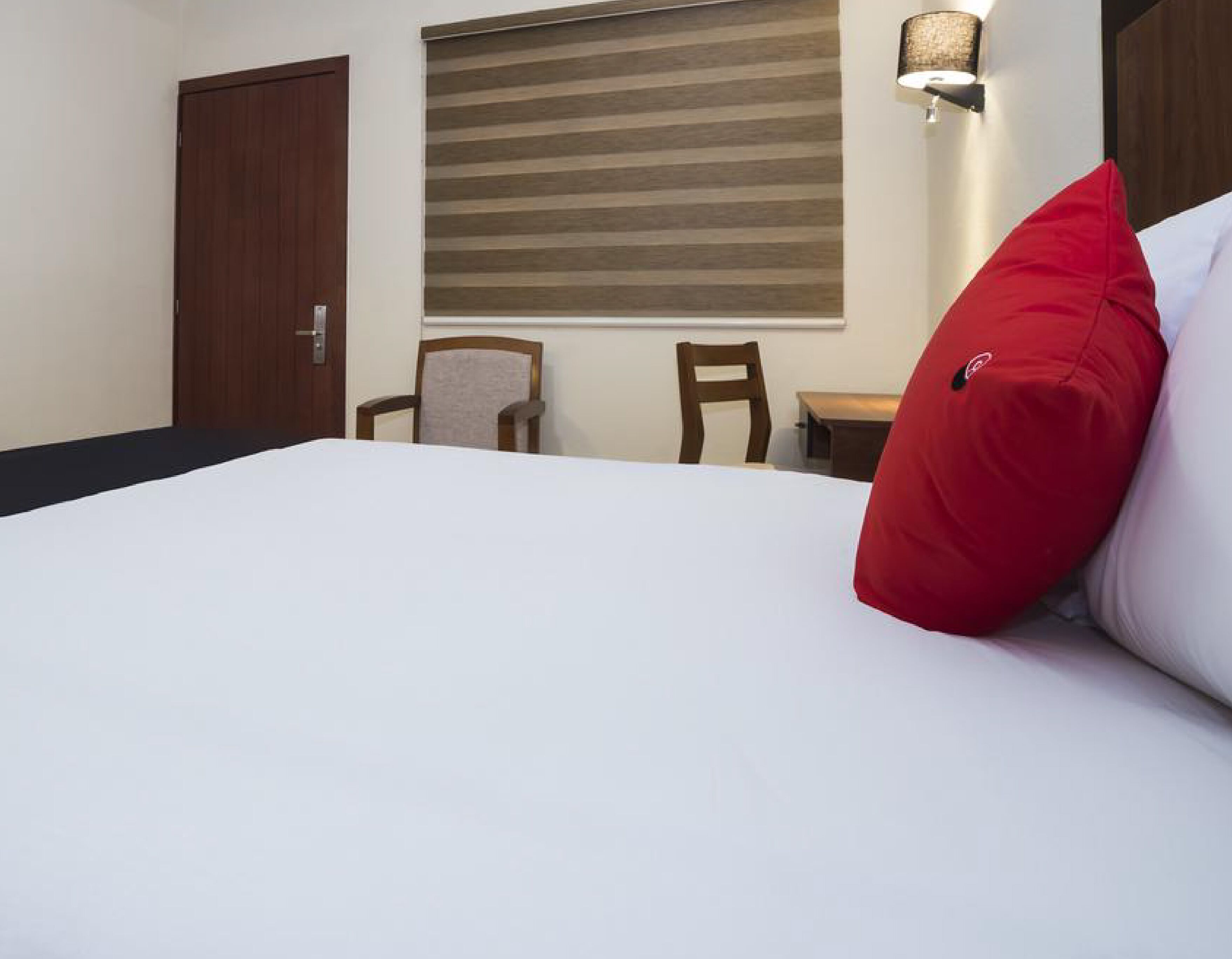 Una habitación de hotel con una cama, dos sillas, una mesita, una ventana con persianas y una lámpara fijada a la pared.