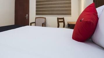 Una habitación de hotel con una cama, dos sillas, una mesita, una ventana con persianas y una lámpara fijada a la pared.