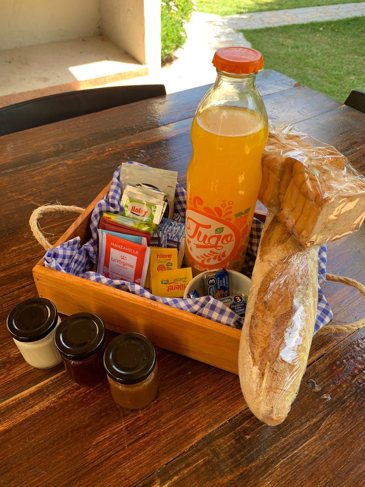 A wooden tray with a bottle of orange juice, bread, condiments, and packaged snacks on a wooden table.