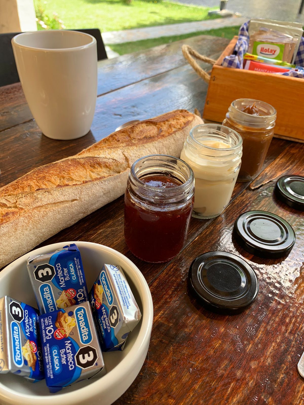 A wooden table with a baguette, jars of jam and butter, and a bowl of cheese sticks.