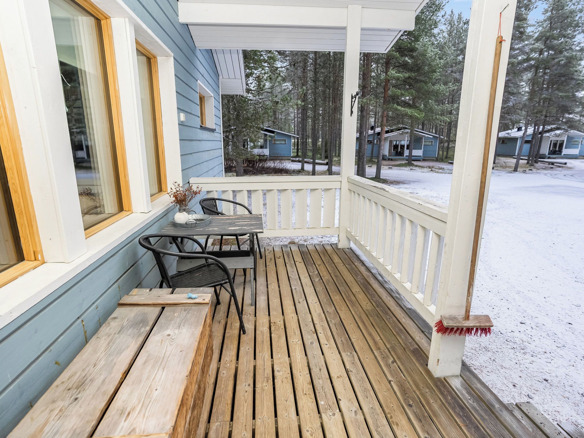 A wooden deck with a table and chairs, a white railing, and a view of a snowy landscape with cabins.
