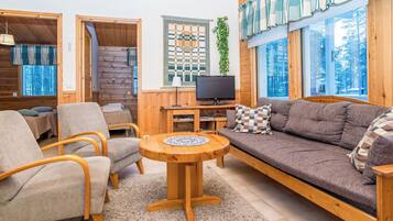 A cozy cabin interior with a sofa, chairs, a TV, and a window with a view of trees.