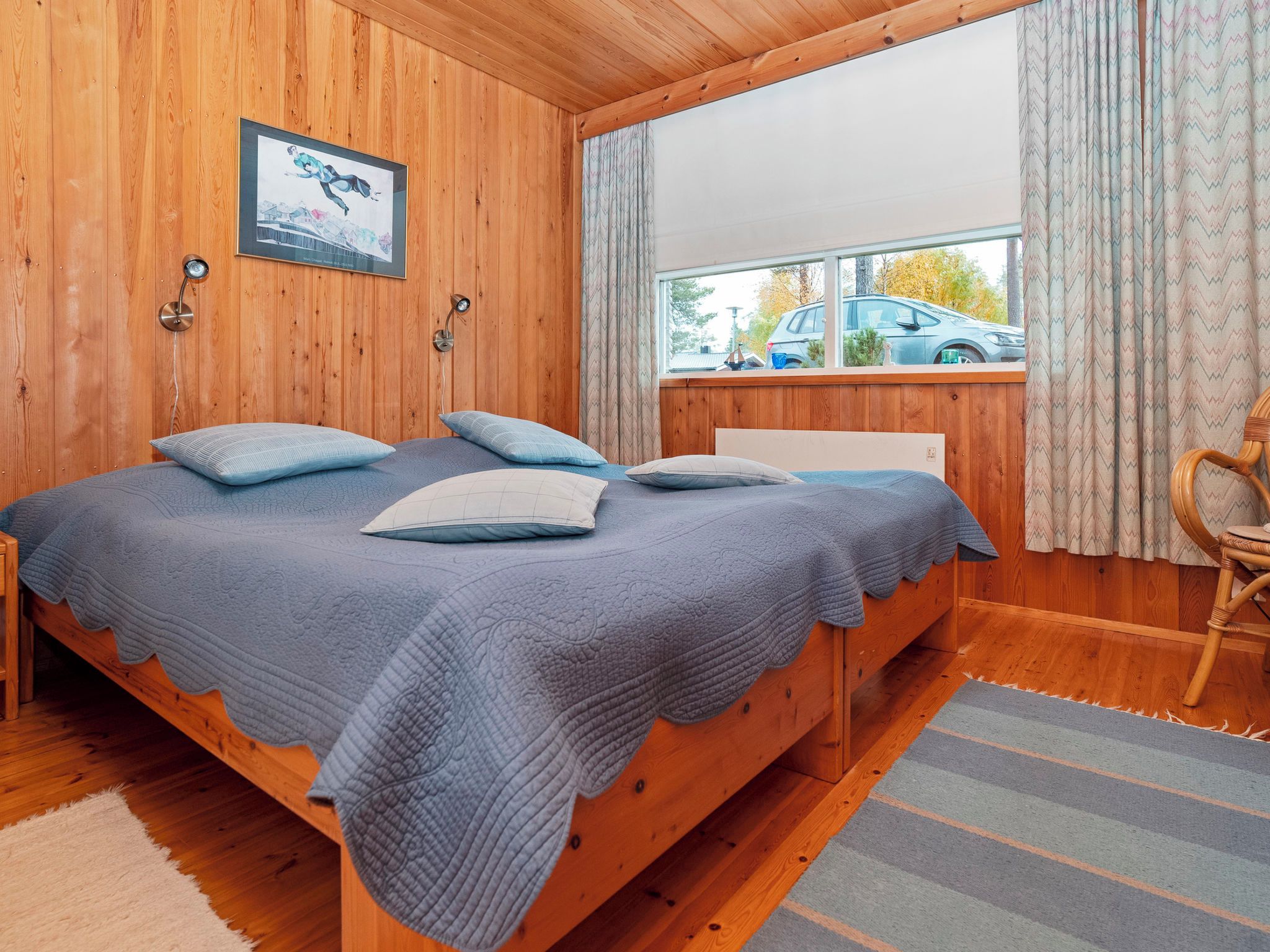 Une chambre dans une cabane en bois, avec un lit, deux lampes fixées au mur, une fenêtre avec des rideaux et une vue sur une voiture à l’extérieur.