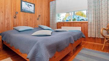 Une chambre dans une cabane en bois, avec un lit, deux lampes fixées au mur, une fenêtre avec des rideaux et une vue sur une voiture à l’extérieur.