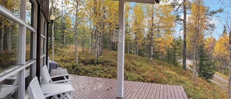 A wooden deck with white chairs, a covered area with a lantern, and a forest view.