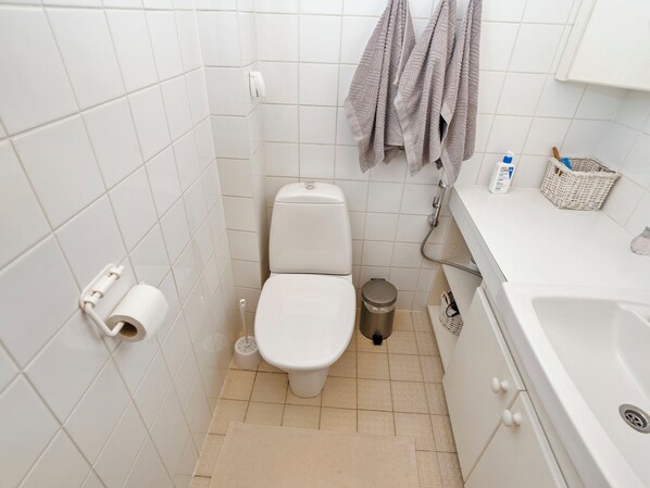 A small, white bathroom with a toilet, a trash can, a toilet brush, a roll of toilet paper, and a showerhead.