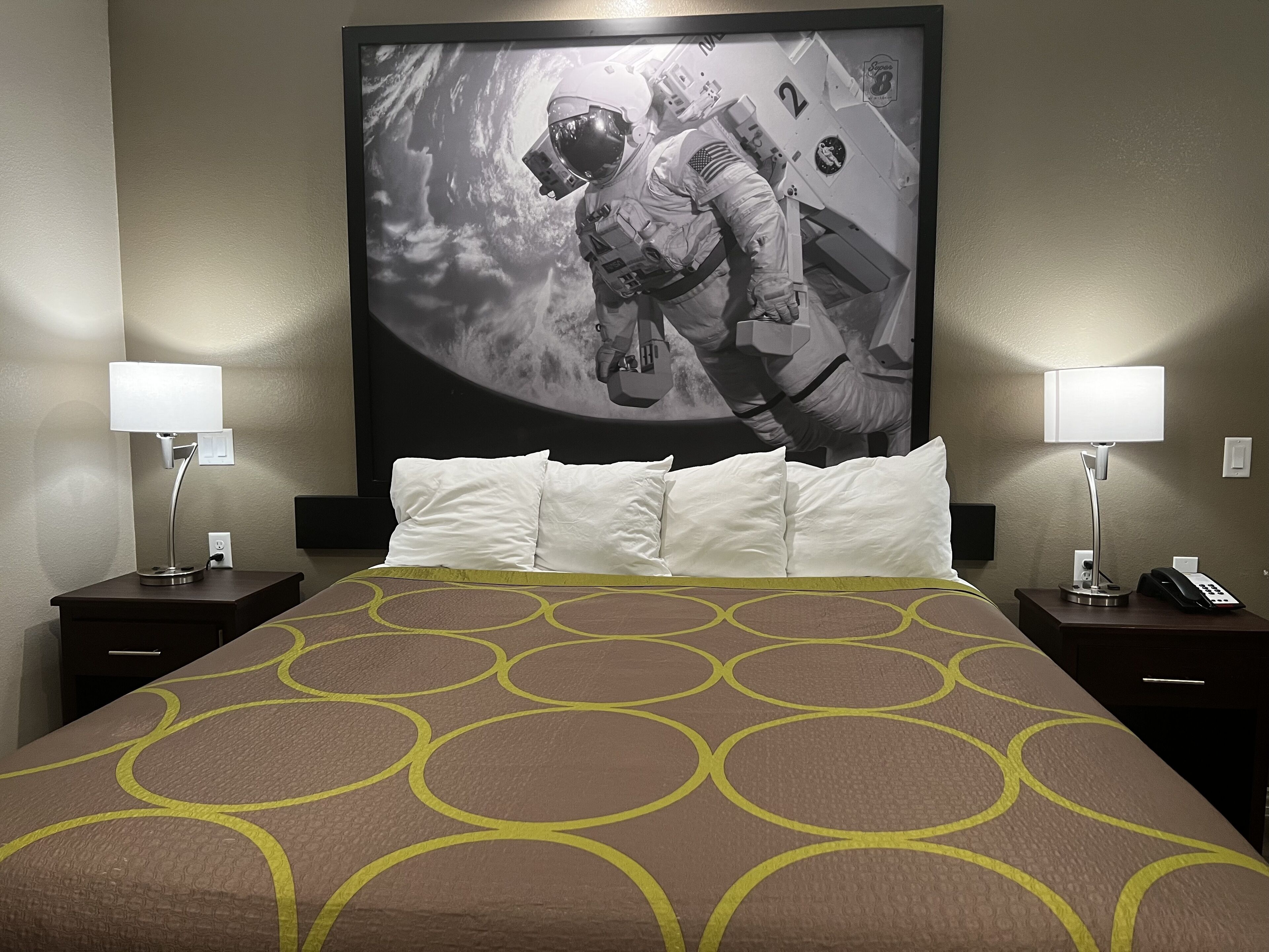 Quarto de hotel com um grande mural de tema espacial na parede, uma cama bem arrumada com colcha estampada, duas mesas de cabeceira com luminárias e telefones, e uma tomada na parede.