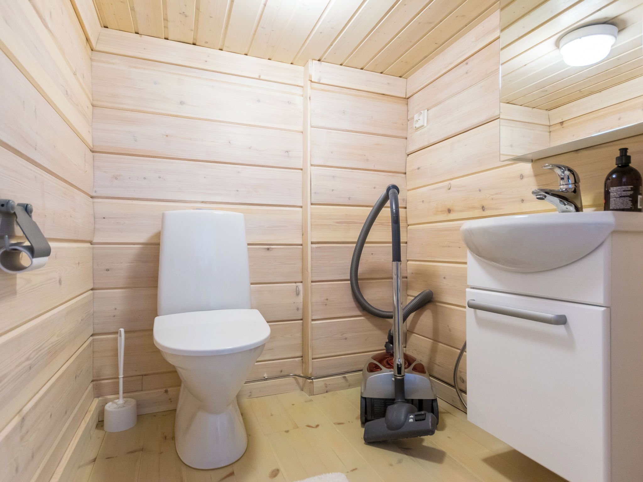 Une petite salle de bain compacte avec des murs en bois, des toilettes blanches, un lavabo avec un miroir et un aspirateur portatif.