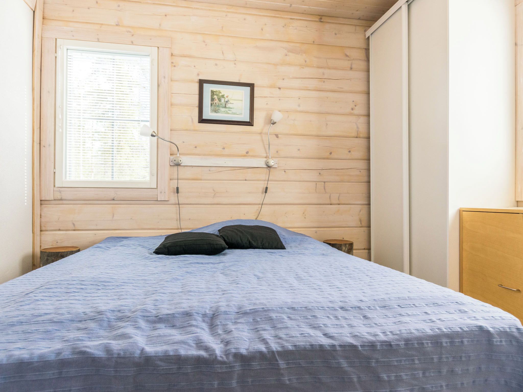 Una habitación con cama, ventana con persianas, un cuadro en la pared y una mesita de noche.