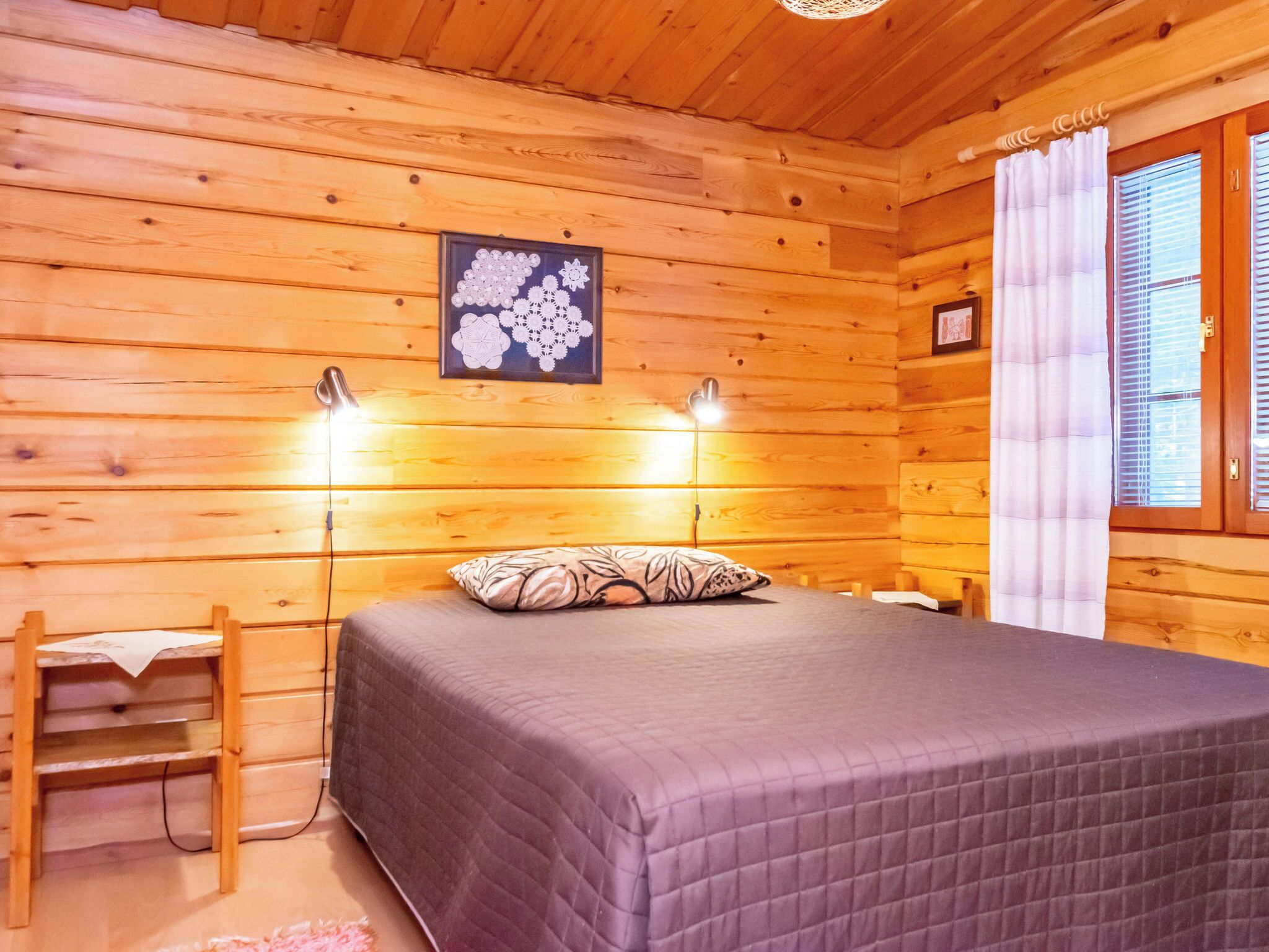 Ett sovrum i en stuga med träpanel, en säng, två sänglampor, ett inramat konstverk med blommotiv och ett fönster med gardiner.