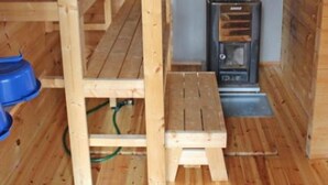 Interior de una cabaña de madera con un estufa de leña, un banco de madera y un contenedor azul.