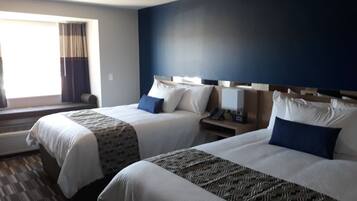 A hotel room with two beds, a large window with curtains, a bedside table with a lamp, and a wall-mounted phone.