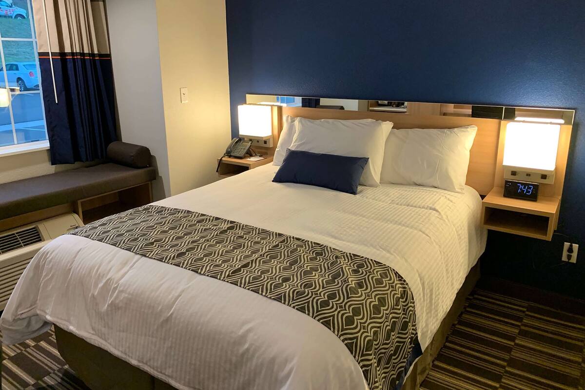 Een hotelkamer met een groot bed, twee nachtkastjes met lampen, een bankje en uitzicht op de straat.