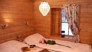 Ein Schlafzimmer einer Holzhütte mit einem Bett, einem Teddybären, einem kleinen Tisch und einem Fenster mit Vorhängen.