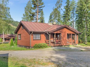 A wooden cabin with a porch, surrounded by trees and a gravel driveway.