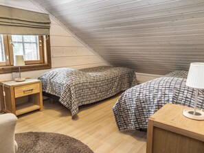 A small, sloped-roofed room with two beds, a nightstand, a lamp, and a window with blinds.