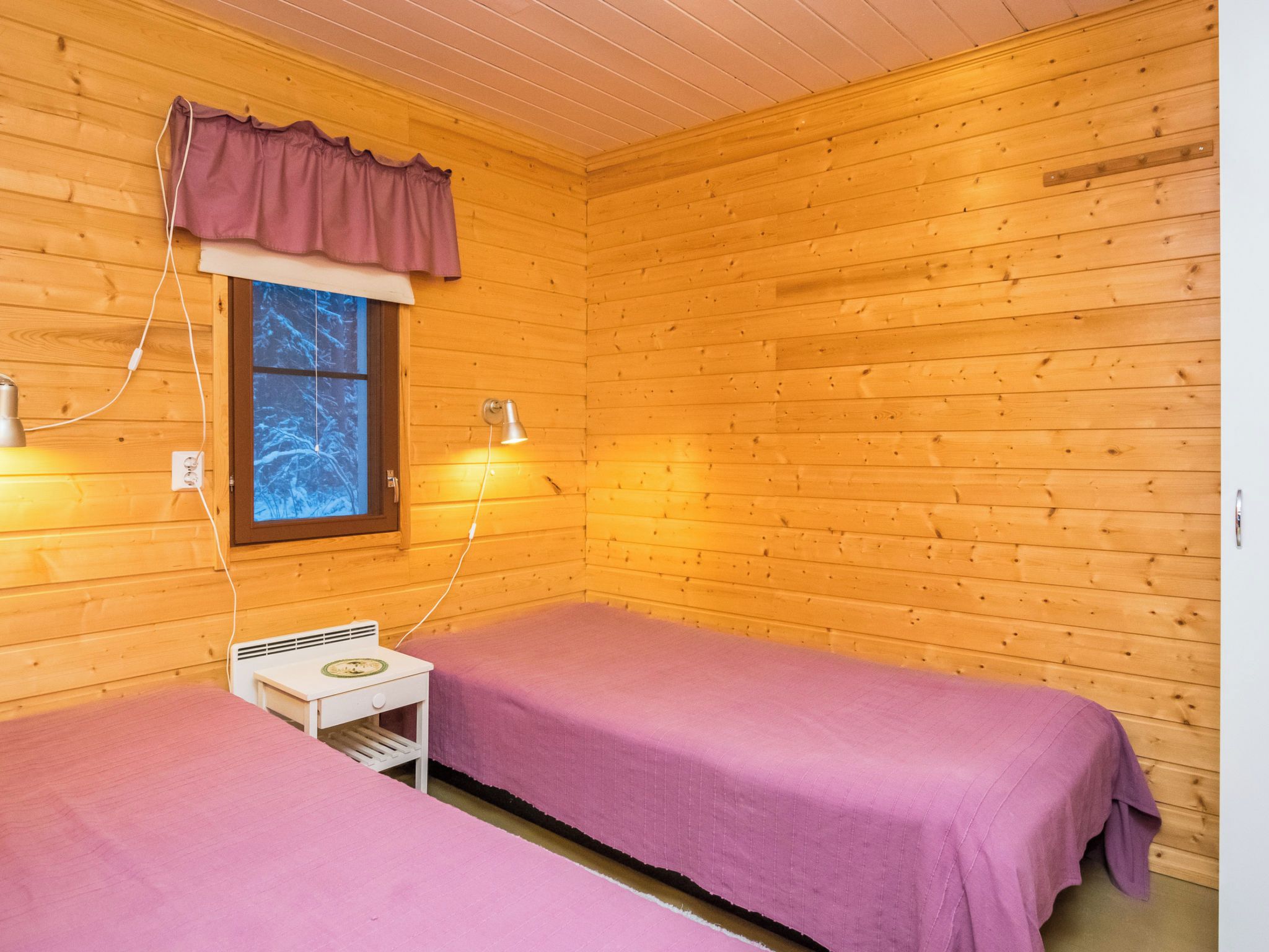 Una cabaña con dos camas, una ventana con vistas a árboles cubiertos de nieve, una mesita con una lámpara y un enchufe.