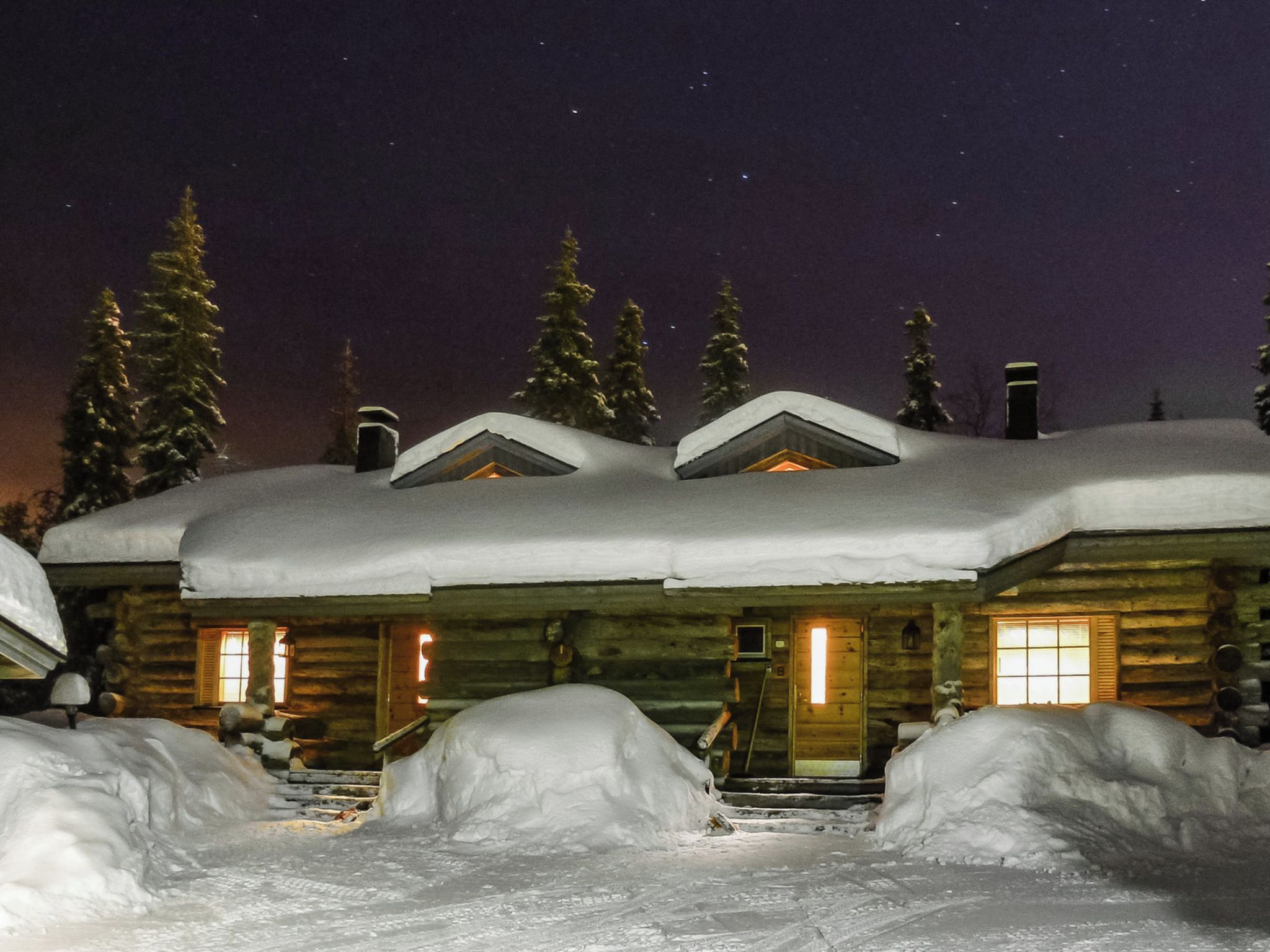 Une cabane en rondins dont le toit est recouvert de neige, et dont la lumière chaleureuse de l’intérieur est visible à travers les fenêtres.