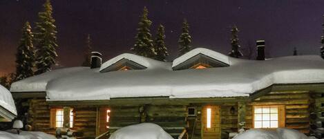 Une cabane en rondins dont le toit est recouvert de neige, et dont la lumière chaleureuse de l’intérieur est visible à travers les fenêtres.