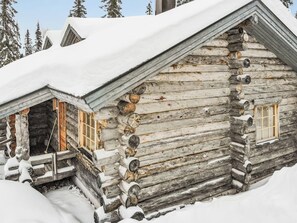 Une cabane en rondins, dont le toit est recouvert de neige et qui possède une cheminée, entourée d’arbres.