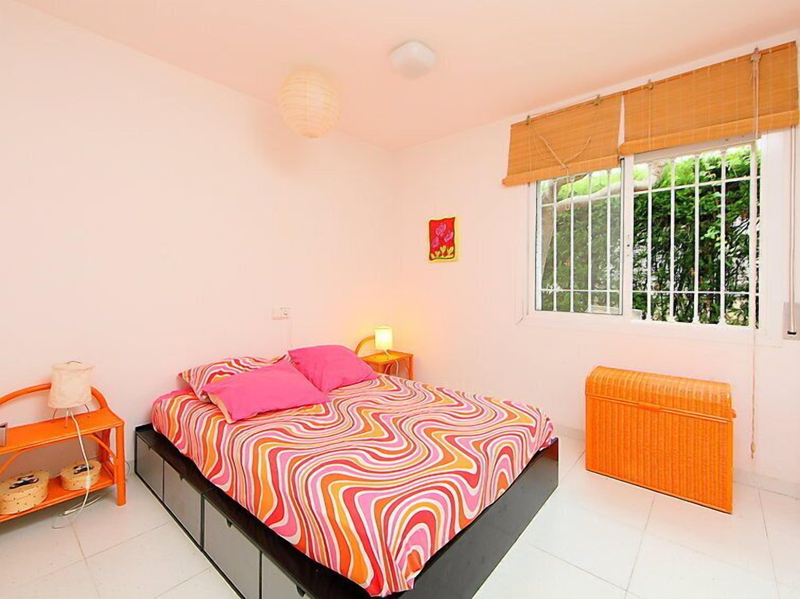 Une chambre à coucher comprenant un lit, une table de chevet, une lampe, une fenêtre avec stores, un radiateur et un élément de décoration murale.