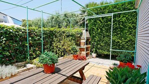 A wooden deck with a brick patio, surrounded by lush greenery and a metal trellis structure.