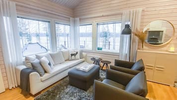 Una sala de estar acogedora con sofá, reposapiés y sillones, una chimenea y ventanas amplias con vistas a árboles.
