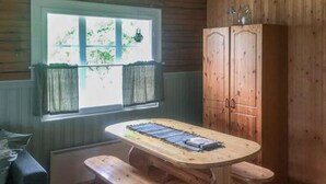 A wooden cabin interior with a dining table and benches, a window with curtains, and a wooden wardrobe.