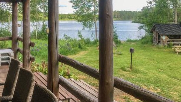 A wooden deck with a chair, overlooking a lake and a small cabin.