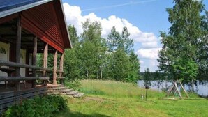 A cabin with a wooden deck and a covered porch, surrounded by trees and a lake.