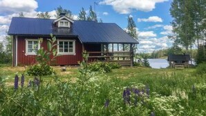 A red wooden cabin with a metal roof, surrounded by greenery and a lake.