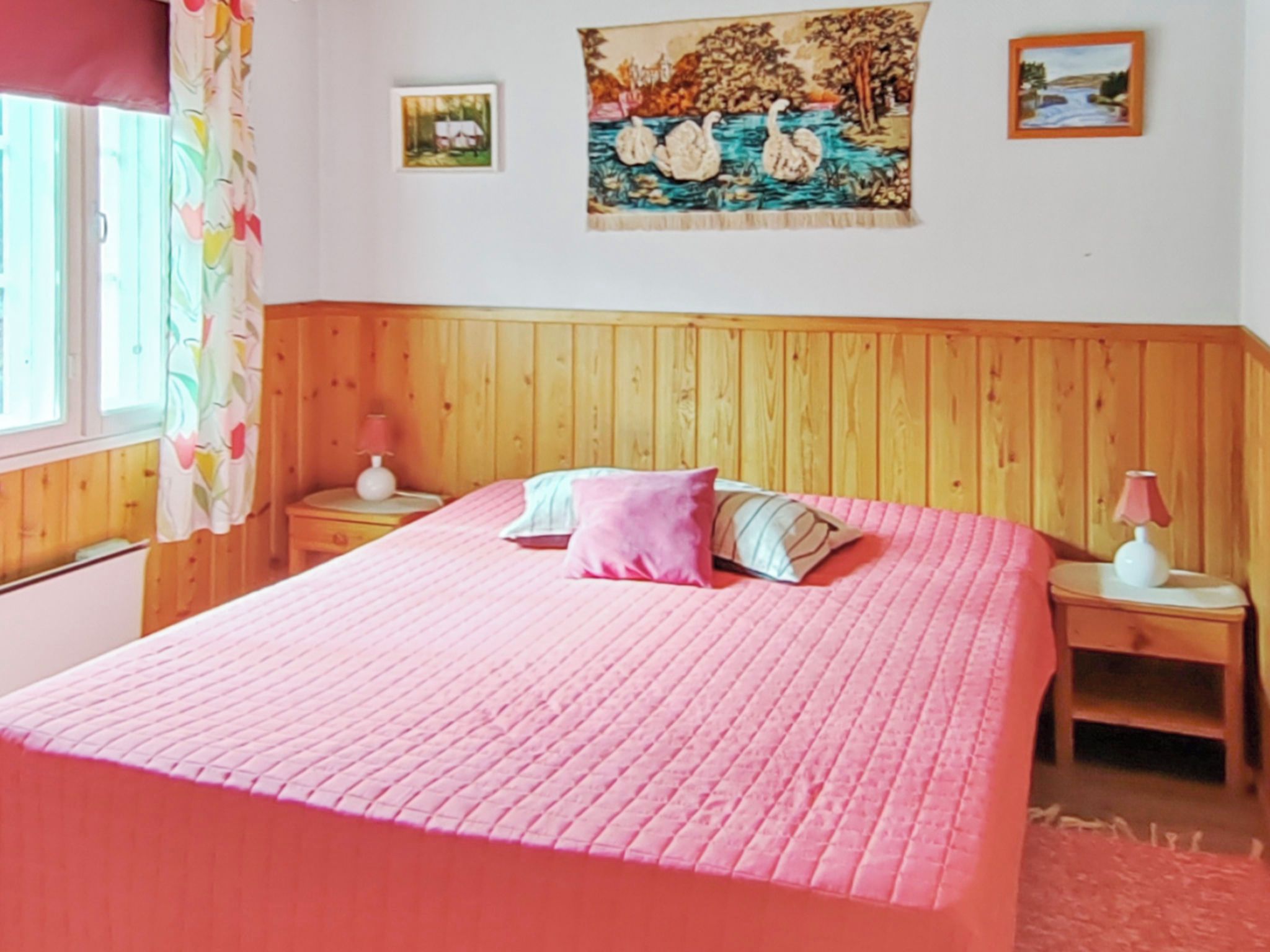 Quarto com cabeceira de madeira, colcha rosa, duas mesas de cabeceira com luminárias e duas imagens emolduradas na parede.