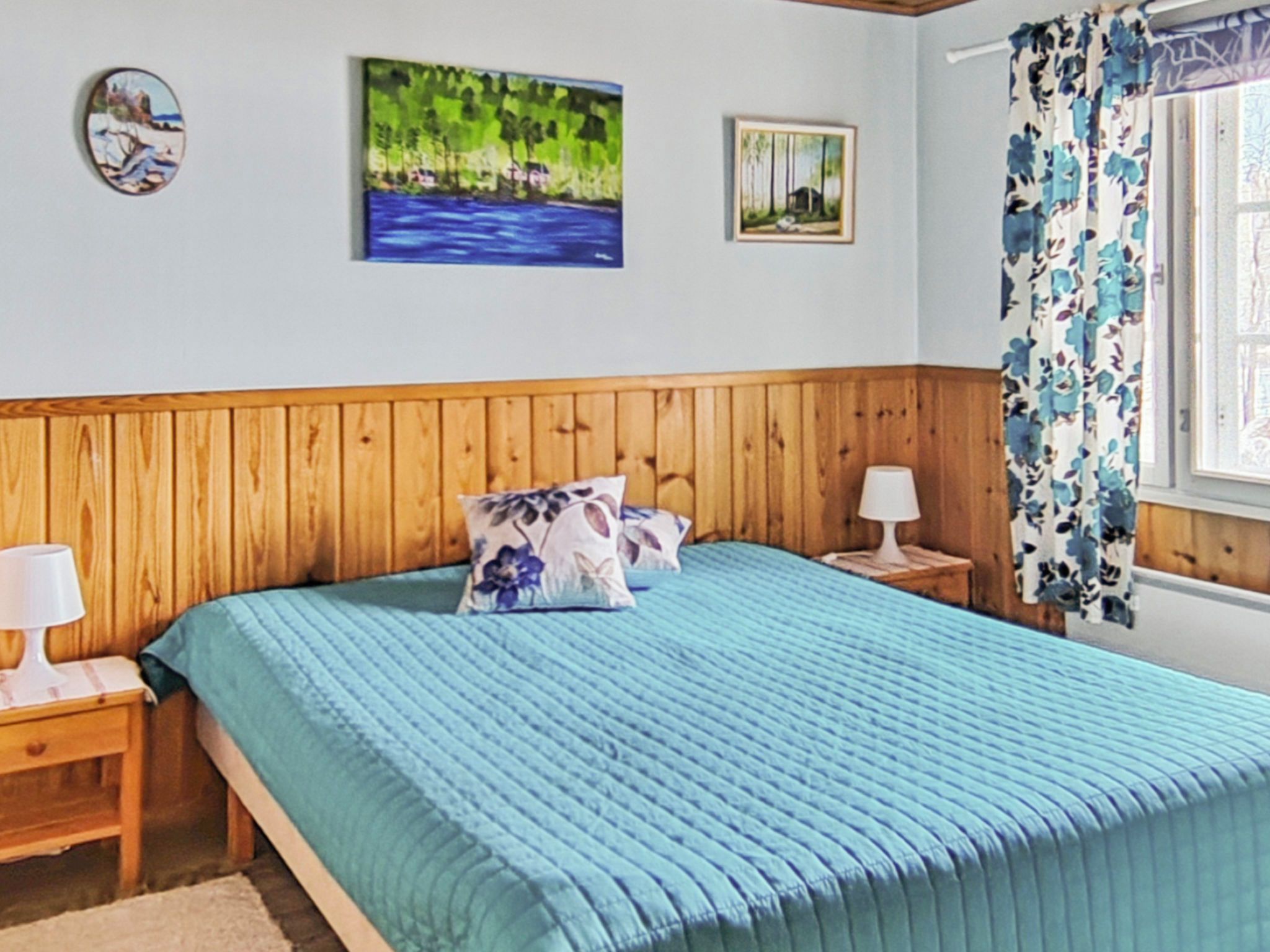 Una camera da letto con una testiera in legno, un letto con biancheria azzurra, due cuscini, un comodino con una lampada e una finestra con tende a fiori.