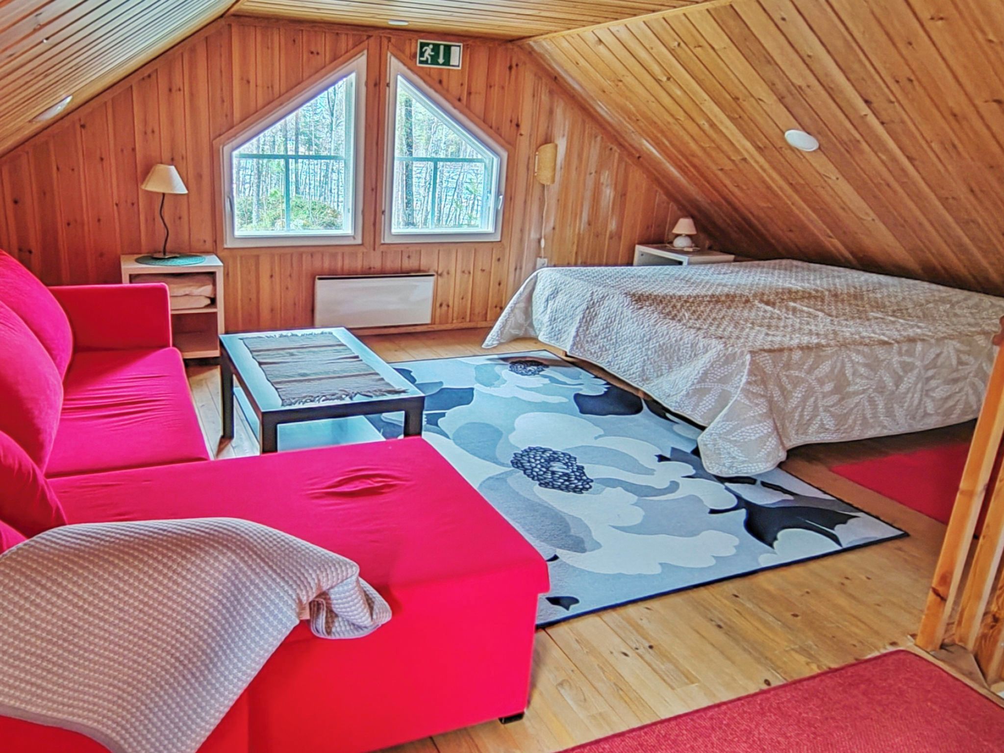 A cozy attic room with a red sofa, a small table, a bed with a patterned bedspread, and two windows with a view of trees.