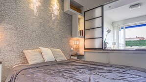 Een moderne slaapkamer met een structuurwand, een groot bed met een grijze deken, een nachtkastje met een lamp, en uitzicht op een groene haag.