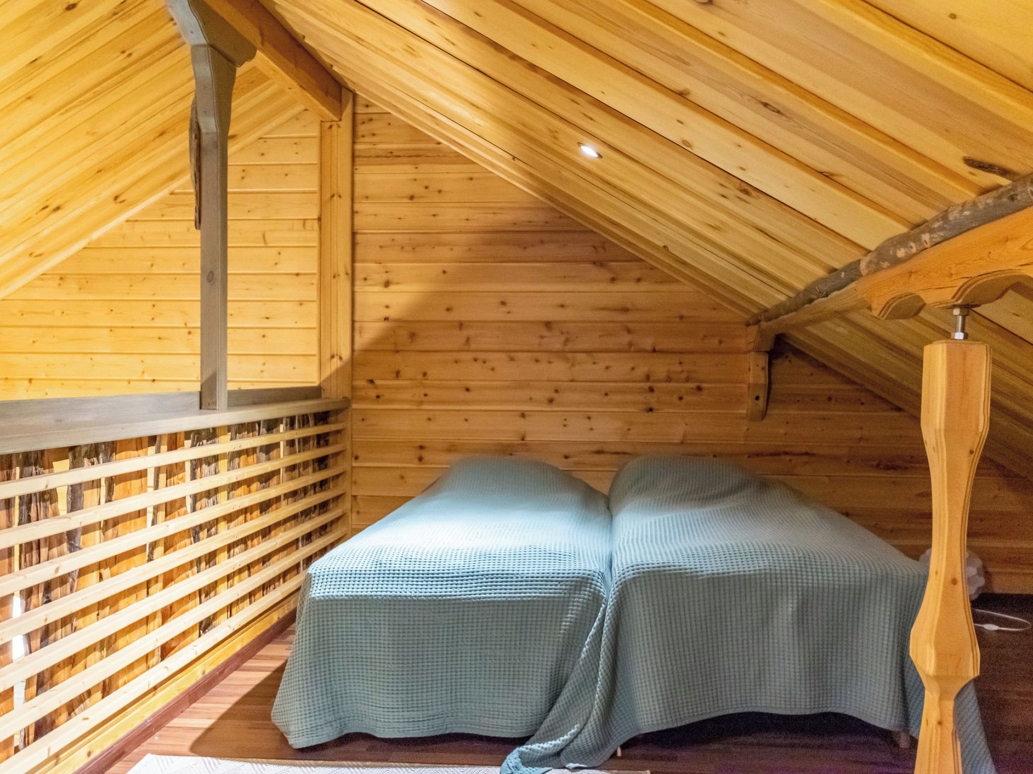 A wooden loft bedroom with a sloped ceiling, a bed with a checkered bedspread, and a built-in shelving unit along the wall.