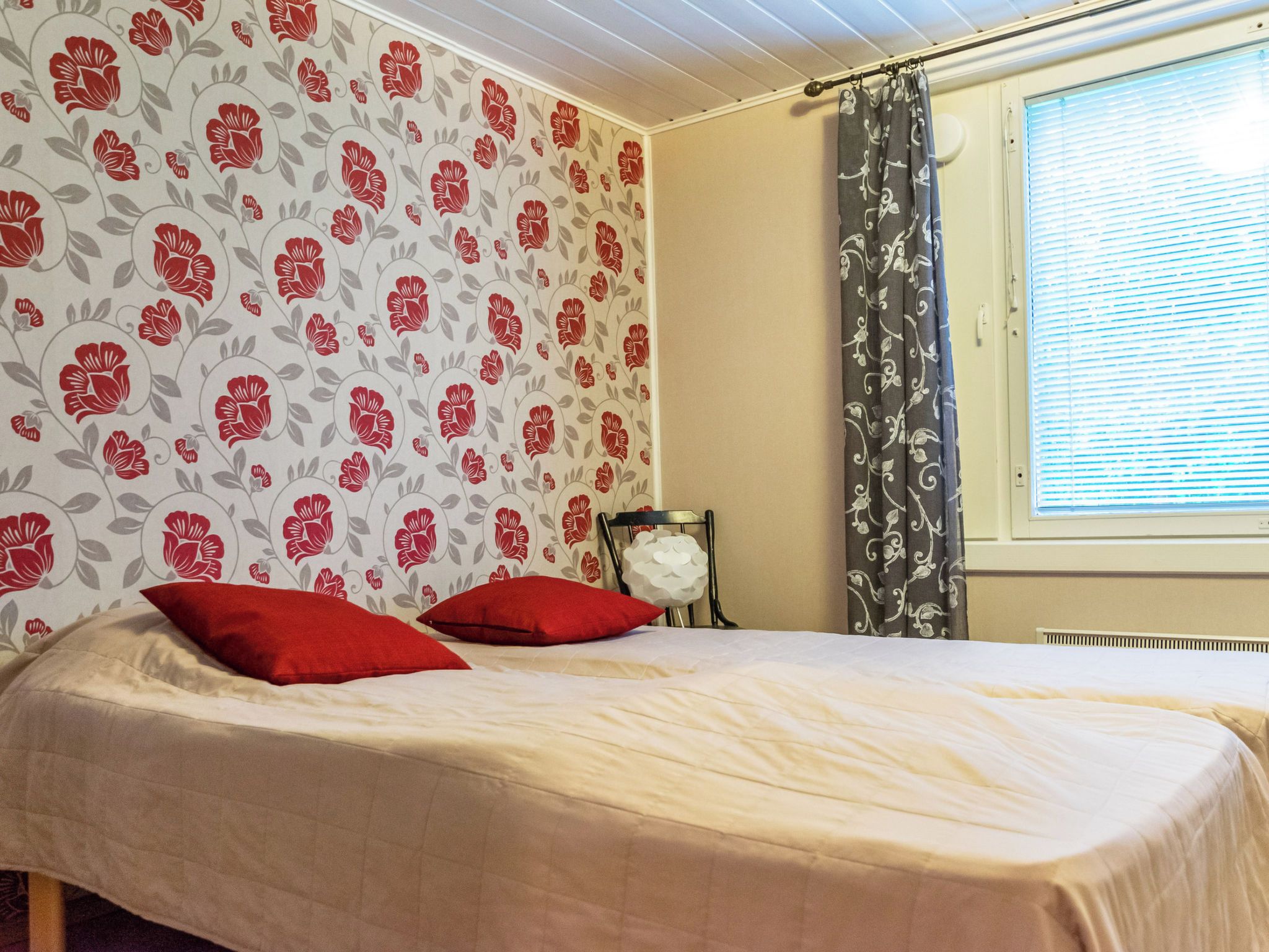 A bedroom with a bed, red pillows, a chair, a window with blinds, and floral wallpaper.