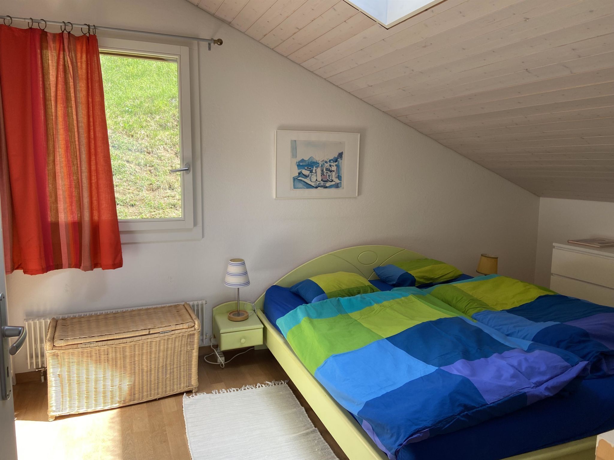 A bedroom with a bed, a window with curtains, a nightstand, a lamp, a wicker basket, a radiator, and a framed picture on the wall.