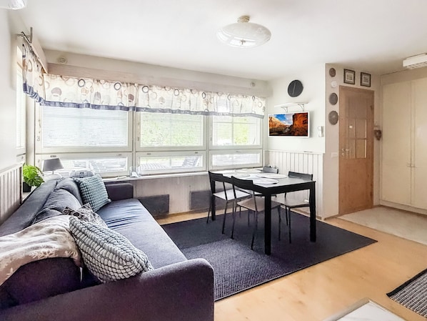 A living room with a sofa, a dining table with chairs, a window with blinds, a radiator, and a door.