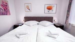 Een tweepersoonsbed met witte lakens, twee kussens en opgevouwen handdoeken. Twee nachtkastjes met lampen staan ernaast. De kamer heeft een neutrale kleurenpalet met kunst aan de muren.